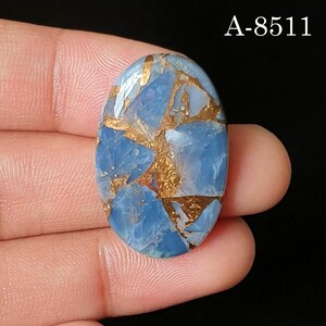 May include: An oval-shaped blue gemstone cabochon featuring a distinct copper-colored matrix pattern throughout the surface. The stone displays a smooth, polished finish with varied shades of light and medium blue. The text A-8511 is visible.