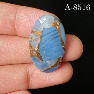 May include: An oval-shaped blue gemstone with metallic copper-colored veining throughout the surface. The stone features a smooth, polished finish and is marked with the text A-8516 in the upper right corner against a black background.