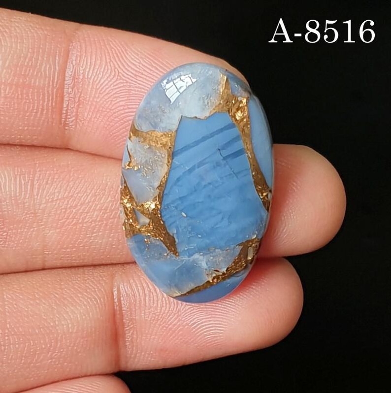 May include: An oval-shaped blue gemstone with metallic copper-colored veining throughout the surface. The stone features a smooth, polished finish and is marked with the text A-8516 in the upper right corner against a black background.