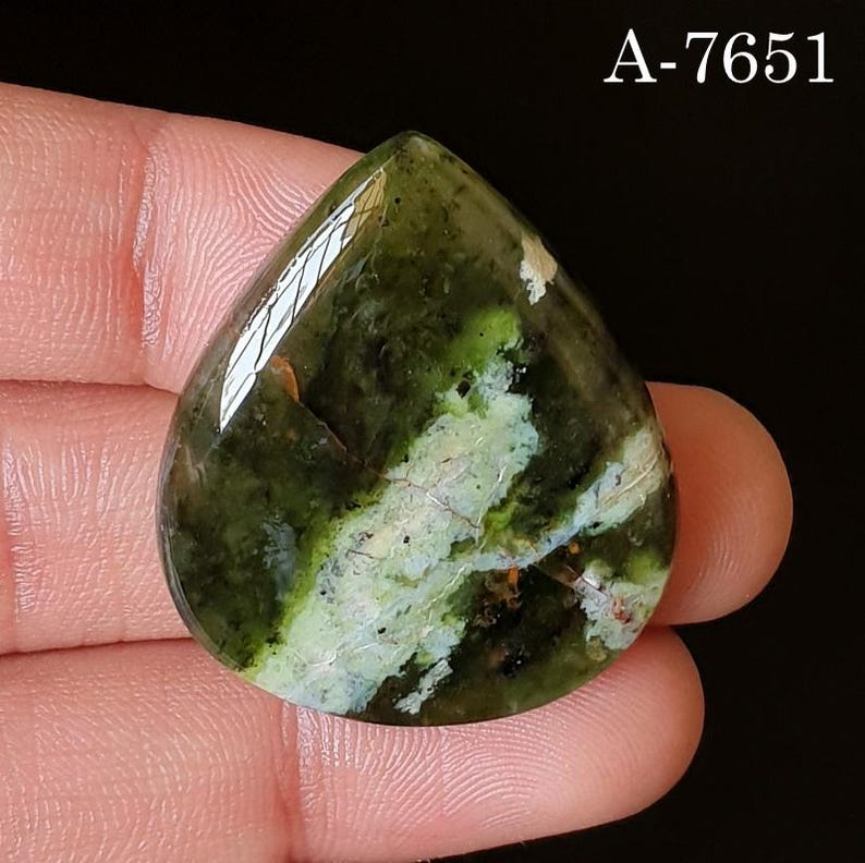 May include: A teardrop-shaped gemstone with a polished surface. The stone features a mix of dark green and light green hues, with a prominent white vein running through the center. The text "A-7651" is visible in the upper right corner. The gemstone is held by a person.