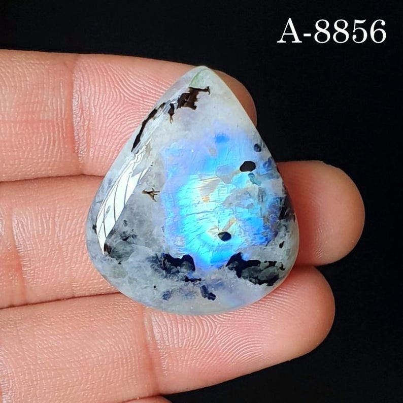 May include: A teardrop-shaped moonstone cabochon featuring a translucent white base with a vibrant blue adularescence in the center and scattered dark mineral inclusions. The stone is marked with the text A-8856 in the upper right corner.
