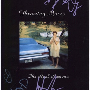 Può includere: Fotografia vintage con bordo nero con il testo "Throwing Muses" e "The Real Ramona". L'immagine mostra una donna vicino a un'auto blu classica. La foto è firmata con inchiostro viola.