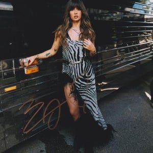 May include: A sleeveless midi dress featuring a black and white zebra print pattern, a black belt with a silver-toned metal buckle at the waist, and a black feathered trim along the asymmetrical hemline, worn with a layered silver chain necklace.