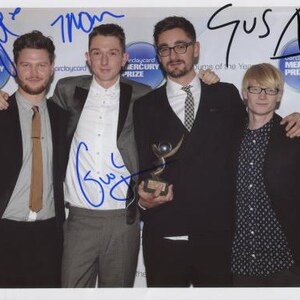 May include: Four men stand together, smiling, holding an award. The award is a silver trophy with a globe on top. The men are wearing suits and ties. The text "Mercury Prize" is visible on the award.