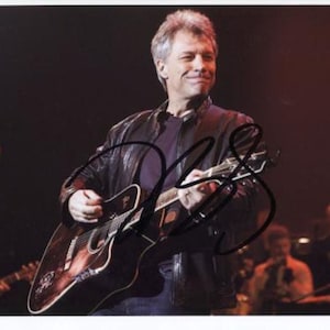 May include: A man in a black leather jacket plays a brown acoustic guitar on stage. He is wearing a black shirt and has short, gray hair. The guitar has a black pickguard and a white pick-up.