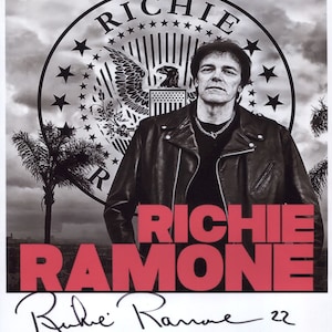 May include: Black and white photograph of Richie Ramone, a musician, wearing a leather jacket. He is standing in front of a large circular graphic with the word "RICHIE" in large letters. The graphic also features a bald eagle and stars. The text "RICHIE RAMONE" is in large red letters below the photo. The photo is signed "Richie Ramone 22".