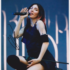 May include: A photograph of a person on stage holding a microphone. The person is wearing a dark blue outfit and black thigh-high socks. The background is a blurred blue with white vertical lines. The person is singing.