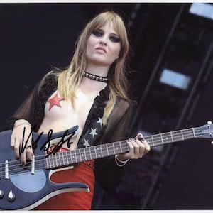 Puede incluir: Una mujer con el pelo largo y rubio toca un bajo eléctrico negro. Lleva una blusa negra transparente con una estrella roja y un collar negro. El bajo tiene una golpeador blanco y una pastilla plateada.