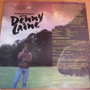 Pode incluir: Capa de álbum de vinil vintage para "hometown girls" de Denny Laine. A capa apresenta uma foto de Denny Laine com uma jaqueta vermelha e jeans, com o título do álbum em letras grandes e estilizadas. O fundo é um céu nublado e um campo gramado.