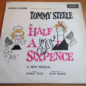 May include: A white vinyl record cover with a pink and black illustration of a man and woman holding half a sixpence. The title of the musical is "Half a Sixpence" and the cover features the name "Tommy Steele" in large black letters. The text "An Original Cast Recording" is printed in black at the top of the cover.