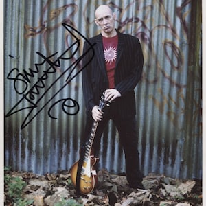May include: A black and white photograph of a man holding a guitar. He is wearing a black suit jacket over a red shirt with a white star design. The man is standing in front of a corrugated metal wall. The photo is signed "Simon Townshend (The Who)".