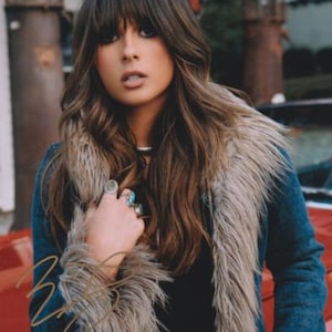 May include: A dark blue denim jacket featuring a prominent beige faux fur collar and trim. The jacket is worn over a black top and is accessorized with multiple silver rings on the fingers. A gold-colored signature is visible on the lower left.