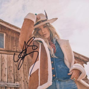 May include: A woman wearing a tan cowboy hat with a feather, a brown suede coat with white faux fur trim, a denim vest, and blue jeans. A red bandana is around her neck. The image is signed in black ink.