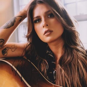 Puede incluir: Una mujer con cabello castaño largo y tatuajes en el brazo sostiene una guitarra acústica marrón. Está mirando a la cámara. La guitarra tiene una tapa marrón claro y un cuerpo marrón más oscuro.