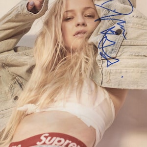 May include: A blonde woman wearing a white crop top, blue jeans, and a red "Supreme" waistband. She is wearing a light blue denim jacket and has her hair pulled back. The woman is looking at the camera and has a slight smile on her face.