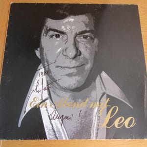 Pode incluir: Capa de álbum de vinil vintage com um retrato em tons de cinza de um homem. O título do álbum, "Ein Abend mit Leo", está em letras douradas. A capa inclui uma assinatura e o ano de 1984.