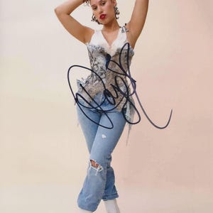 May include: A woman in a white and blue patterned corset top, light blue ripped jeans, and white platform boots. Her hair is styled up, and she is posing with her arms above her head. The image has an autograph in black ink.