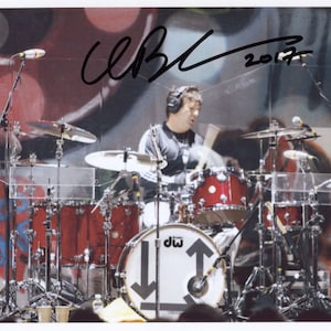 May include: A drummer plays a drum kit with a DW logo on the bass drum. The drummer is wearing headphones and a black shirt. The background is a graffiti wall with the signature "C.B. 2017".