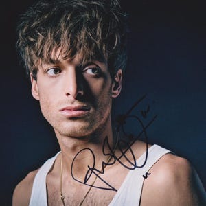 Puede incluir: Retrato de una persona con piel clara, cabello castaño y una camiseta sin mangas blanca. La persona mira hacia un lado con expresión seria. Una cadena dorada es visible alrededor del cuello. La imagen está firmada con tinta negra.