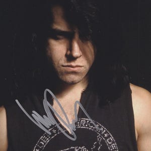May include: A portrait of a person with long, dark hair and a serious expression. They are wearing a black sleeveless shirt with a circular graphic design and a signature. The image is in a dark setting.