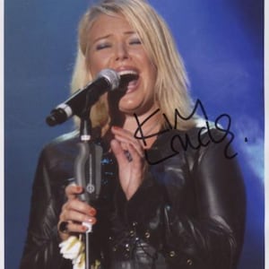 Puede incluir: Una mujer con cabello rubio canta en un micrófono mientras lleva una chaqueta de cuero negra. La firma "Kim Wilde" está escrita en tinta negra en la imagen.