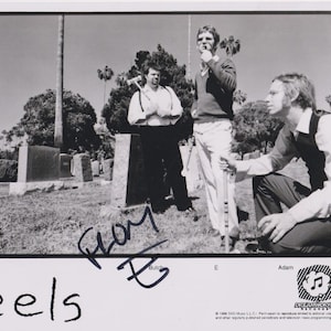 May include: A black and white photograph of the band Eels in a graveyard. Three men are posing near gravestones, one holding a tool. The band's name, "eels", is written at the bottom left, with signatures and names of the band members.