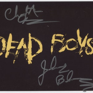 Puede incluir: Fondo negro con las palabras "DEAD BOYS" en pintura dorada. Las firmas de los miembros de la banda están en tinta gris arriba y abajo del nombre de la banda.