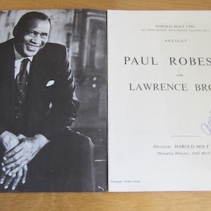 May include: A monochrome photograph of Paul Robeson, smartly dressed in a suit, alongside a printed page. The page reads "PAUL ROBESON with LAWRENCE BROWN" and features a handwritten signature. The items are on a light surface.