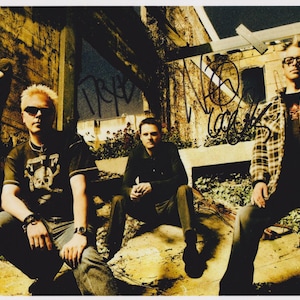 May include: A photograph of a rock band posing outdoors. The band members are wearing sunglasses and casual clothing. The background features graffiti and a weathered building. The image has a vintage, sepia-toned aesthetic.