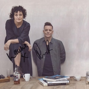 May include: Two people, a woman and a man, are standing in front of a white wall. The woman is wearing a black dress and the man is wearing a grey shirt. They are both looking at the camera. The woman has signed her name "Leanne" and the man has signed his name "Mark".