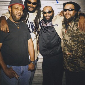 May include: A group of four men posing for a photo, all wearing sunglasses. One man is wearing a black t-shirt and jeans, while another is wearing a camouflage jacket. The photo has signatures in blue and black ink.