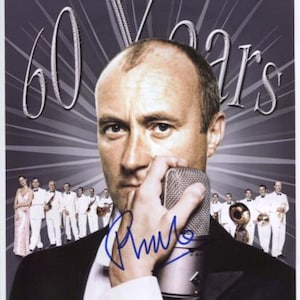 May include: A man with a microphone in front of him, holding it with his right hand. He is wearing a black suit and a white shirt. The background is a silver and black design with the text "60 Years" in large white letters. The man's signature is visible in blue ink.