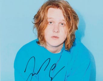 Op de afbeelding: Een portret van een persoon met halflang lichtbruin haar in een felblauw sweatshirt met ronde hals. De afbeelding bevat een handtekening in zwarte inkt op de voorkant van het sweatshirt tegen een effen lichtblauwe achtergrond.