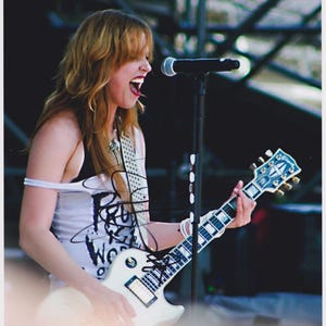 Puede incluir: Foto de una músico tocando una guitarra eléctrica blanca en el escenario. La músico lleva una camiseta sin mangas blanca con texto negro y una correa de guitarra con tachuelas. Un micrófono está colocado frente a la músico.