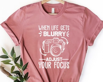 May include: A dusty rose t-shirt with a white graphic of a camera and the text "When life gets blurry, adjust your focus".