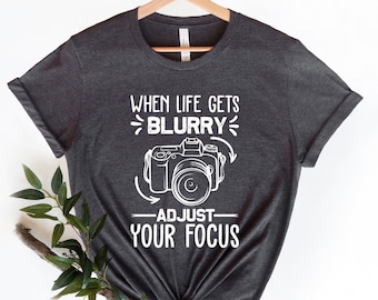 May include: A dark gray t-shirt with a white graphic design. The graphic features a camera with the text "When life gets blurry, adjust your focus".