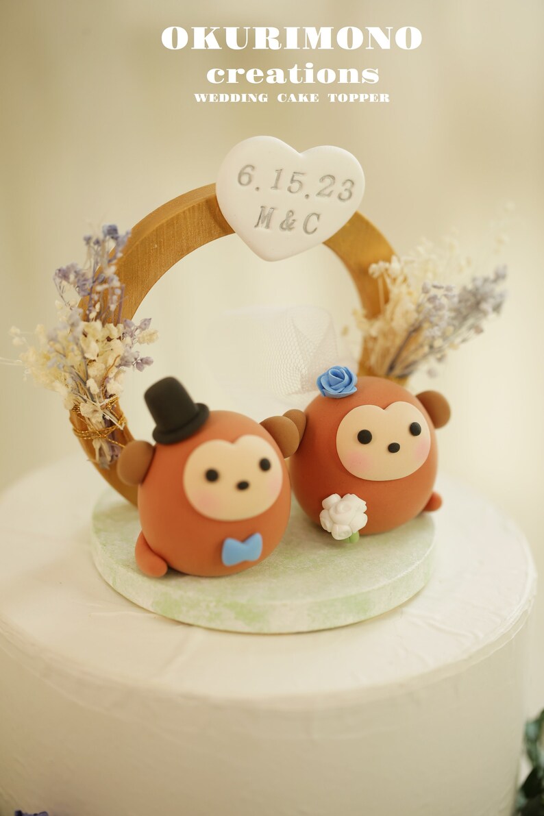 Puede incluir: Un adorno para pastel de boda con dos figuritas de mono marrones con detalles azules. Los monos est&aacute;n de pie bajo un anillo de madera con un letrero en forma de coraz&oacute;n que dice "6.15.23 M&C".