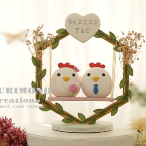 May include: White ceramic chicken figurines on a swing, with a heart-shaped sign that reads "04.23.22 T&C", surrounded by greenery. The swing is part of a wooden hexagonal frame with a leafy vine design. This is a wedding cake topper.