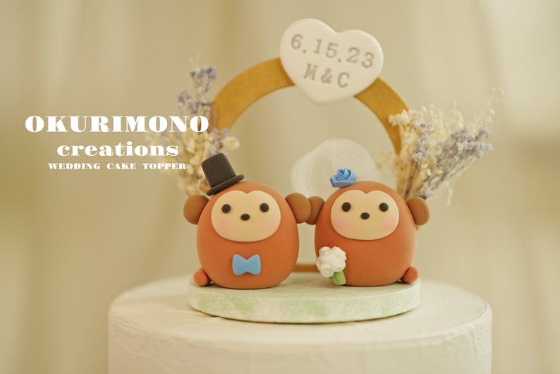 Puede incluir: Un adorno para pastel de boda con dos figuritas de mono marrones que llevan un sombrero de copa y una flor azul. Los monos est&aacute;n de pie sobre una base de pastel blanca con un arco de madera y un cartel en forma de coraz&oacute;n que dice "6.15.23 M&C".