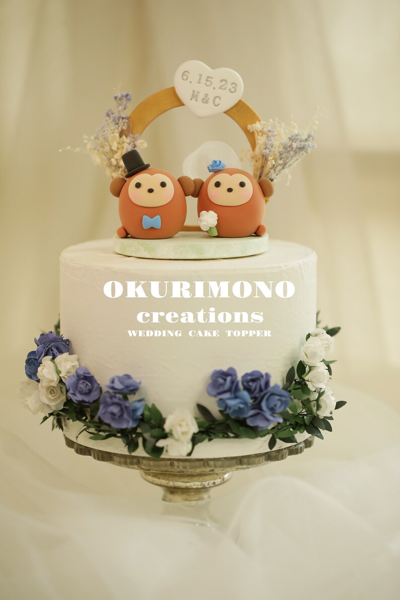 Puede incluir: Un pastel de bodas blanco con un adorno de pastel de pareja de monos marr&oacute;n y blanco. El adorno presenta un arco de madera con la fecha 6.15.23 y las iniciales H&C. El pastel est&aacute; decorado con flores azules y blancas.