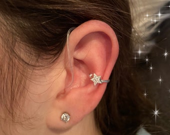May include: A close-up of a person's ear with a silver star-shaped ear cuff with clear rhinestones. The ear cuff is worn on the upper part of the ear, above the lobe. The person also has a small, clear stud earring in their lower ear lobe.