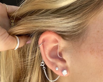May include: A close-up of a person's ear with multiple earrings. The person has a silver chain earring with a small pearl and a square diamond-shaped earring. The person also has a small hoop earring in their upper ear.