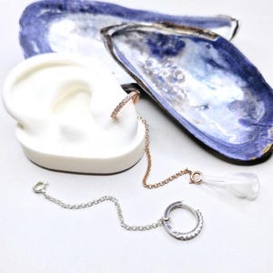 Earrings with a chain and a small hoop. One earring is a small hoop with clear stones, connected by a chain to a larger hoop with clear stones. The other earring is a small hoop with clear stones, connected by a chain to a clear, teardrop-shaped charm. The earrings are displayed on a white ear model and next to blue and white shells. These are used to attach to our holster and helps keep an apple airpod in place