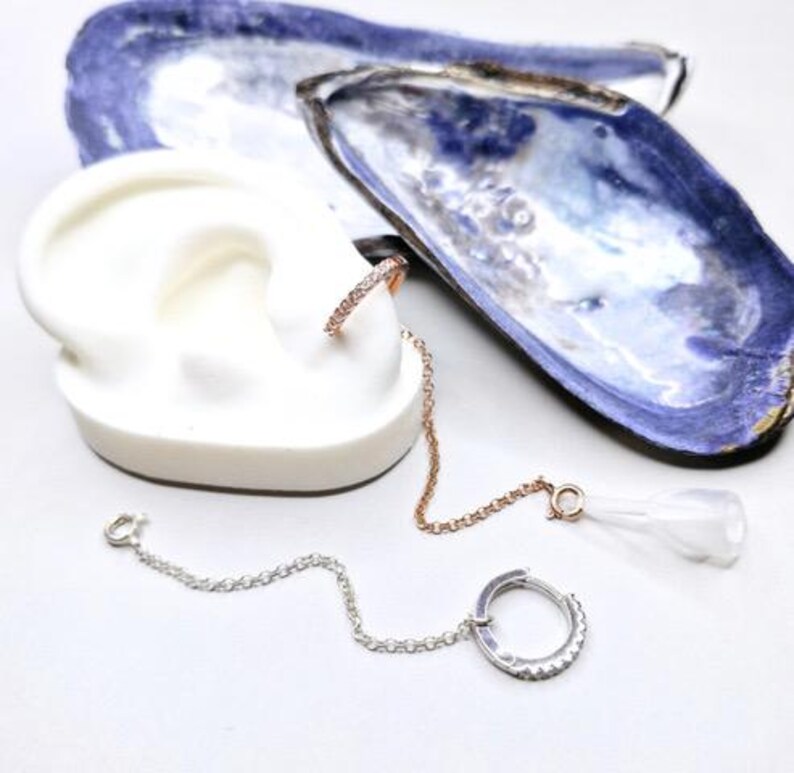 Earrings with a chain and a small hoop. One earring is a small hoop with clear stones, connected by a chain to a larger hoop with clear stones. The other earring is a small hoop with clear stones, connected by a chain to a clear, teardrop-shaped charm. The earrings are displayed on a white ear model and next to blue and white shells. These are used to attach to our holster and helps keep an apple airpod in place