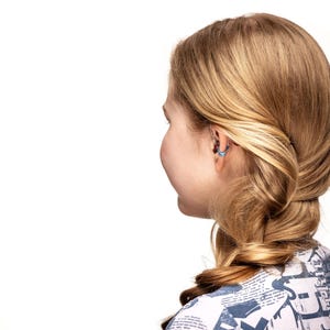May include: A side view of a person with blonde hair styled in a braid. The person is wearing a patterned top and has a small, light blue earring in their ear. The background is white.
