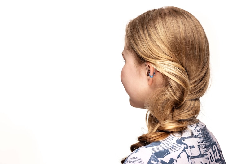 May include: A side view of a person with blonde hair styled in a braid. The person is wearing a patterned top and has a small, light blue earring in their ear. The background is white.