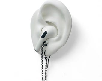 May include: A white ear sculpture with a white AirPod in the ear canal. A black chain earring is attached to the AirPod and dangles down. The background is plain white.