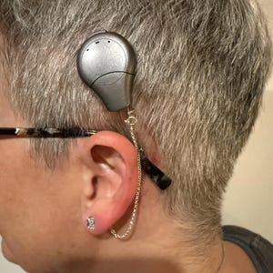 Può includere: Un apparecchio acustico argentato attaccato all'orecchio con una catena. Il dispositivo è collegato a un paio di occhiali. Un piccolo orecchino quadrato e trasparente è visibile nell'orecchio. La persona ha i capelli corti e grigi.
