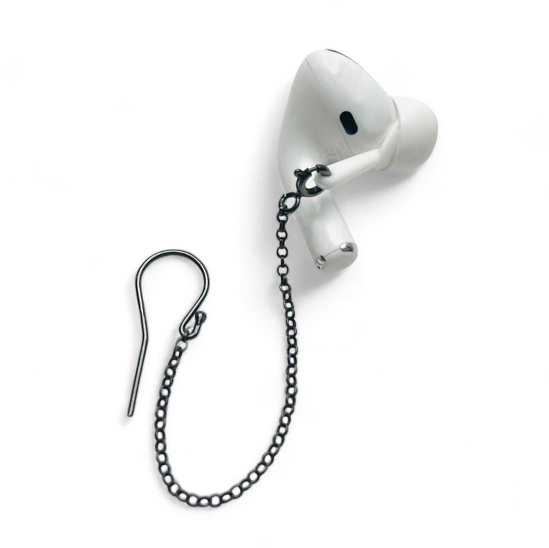 A white wireless earbud with a black chain earring. The earbud has a rounded shape with a black accent. The earring chain is made of small, dark gray links, connecting the earbud to a hook-shaped earring. These are used to prevent loss of airpods it is a lanyard
