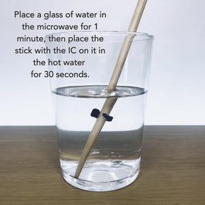May include: A clear glass of water with a wooden stick and a black clip inside. Text on the image reads: Place a glass of water in the microwave for 1 minute, then place the stick with the IC on it in the hot water for 30 seconds.